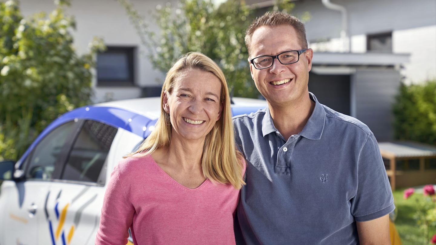 Lächelndes Paar vor Haus und weißem Auto.