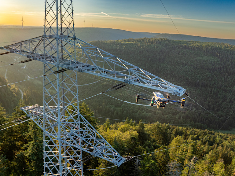 Eine Drohne fliegt an einem Freileitungsmasten entlang.