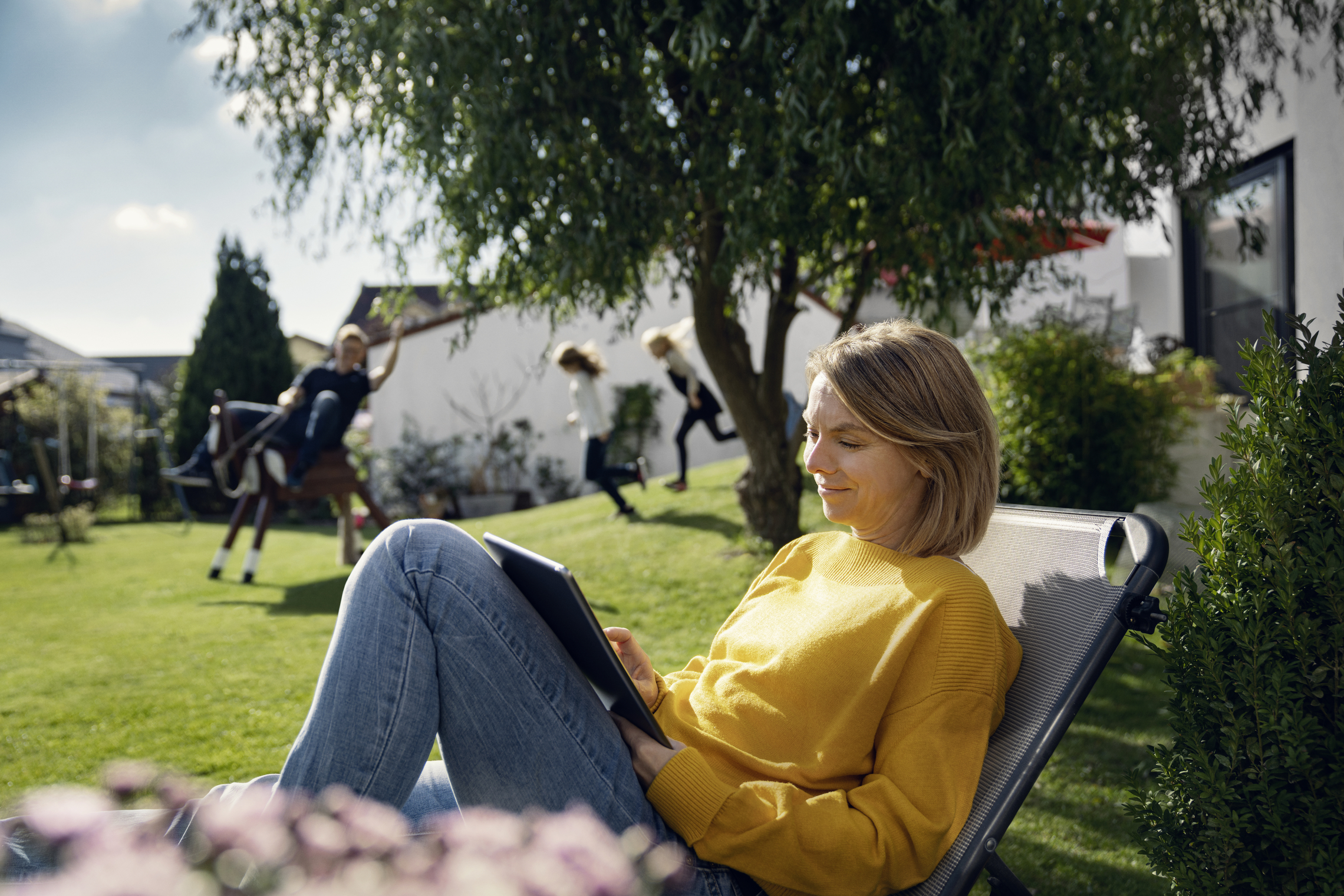 Frau entspannt im Garten, Tablet in der Hand. Kinder spielen im Hintergrund.