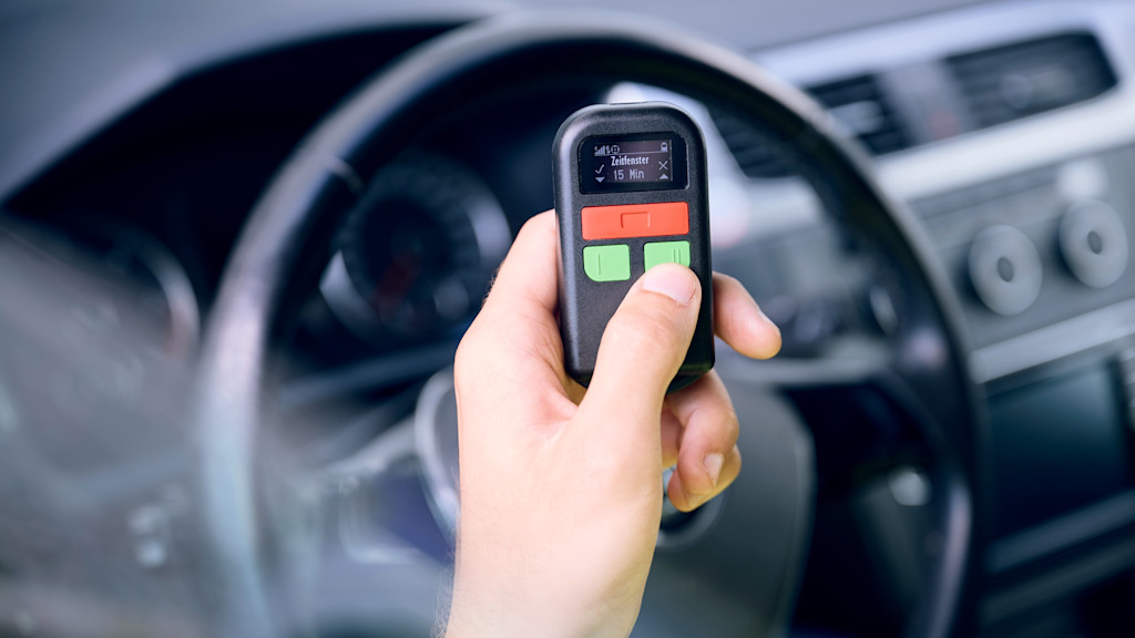 Hand hält schwarzes Gerät mit Anzeige "Zeitfenster: 15 Min" im Auto.