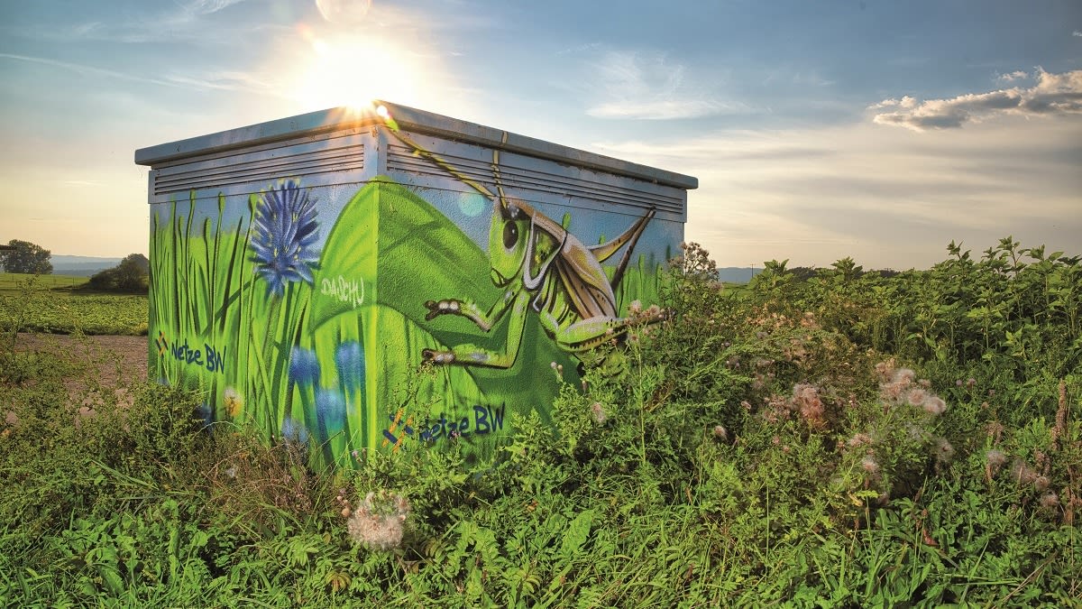 Graffiti an Schaltkasten: Heuschrecke in grüner Landschaft.
