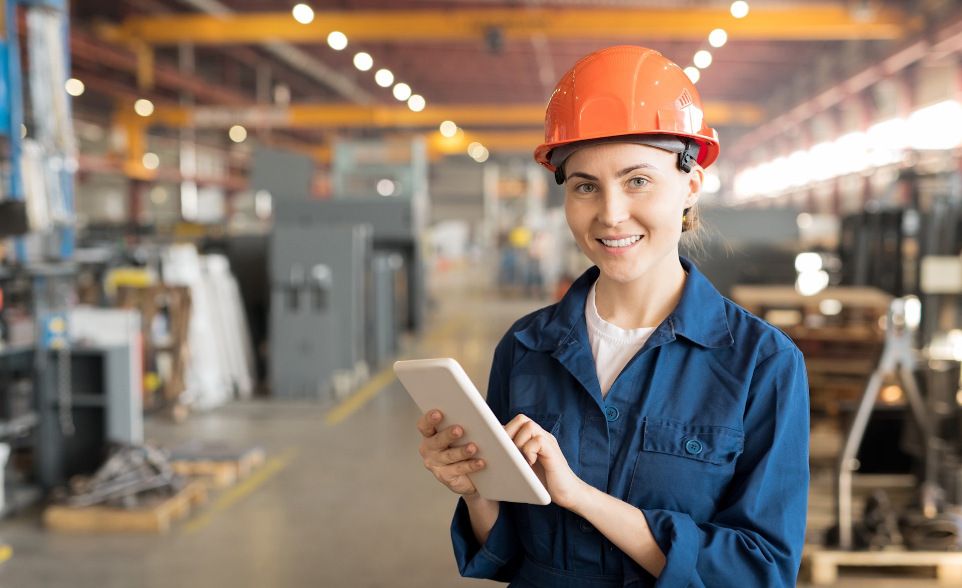 Lächelnde Arbeiterin mit Tablet steht in einer Fabrikhalle.