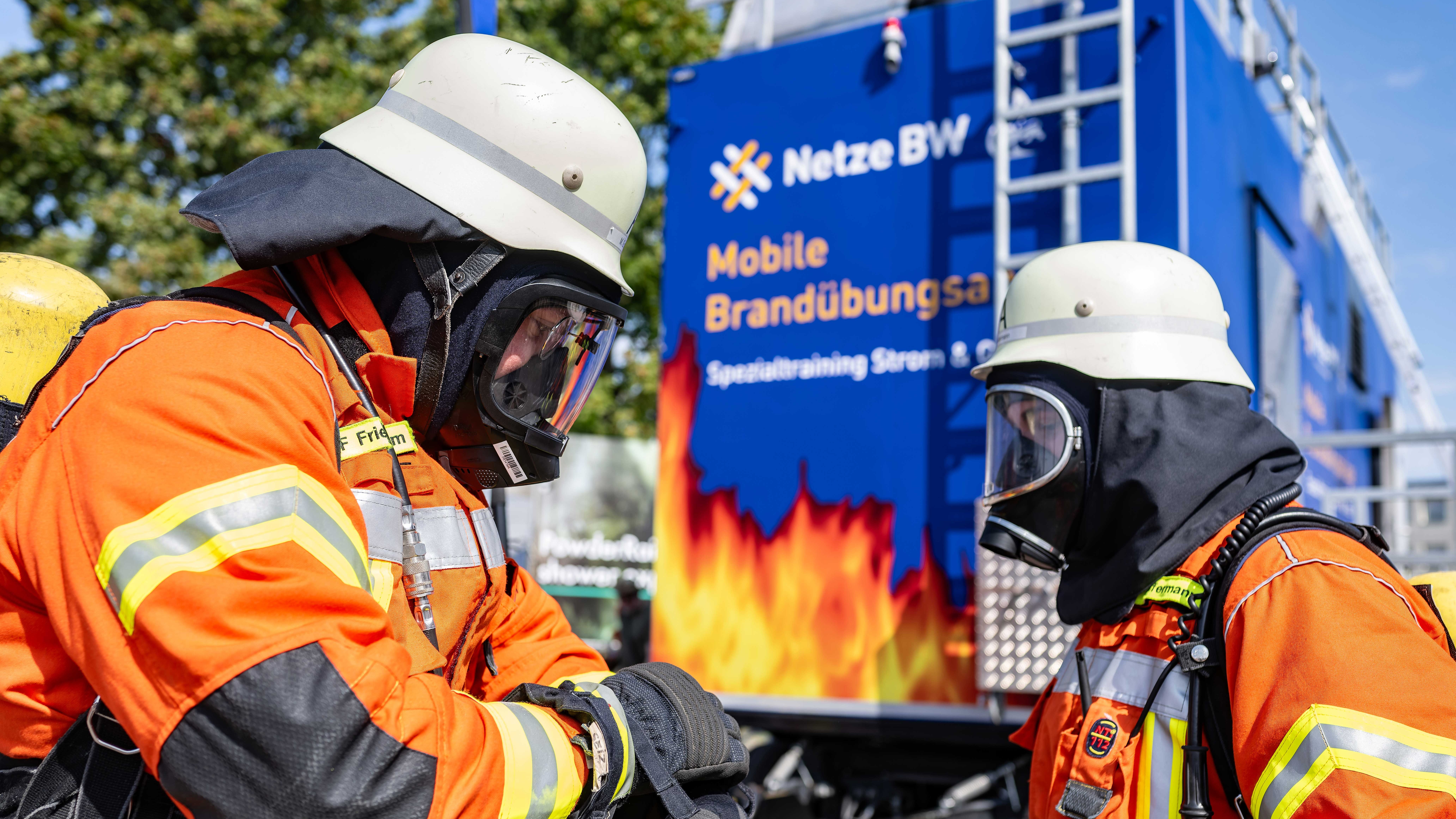 Zwei Feuerwehrleute in Schutzkleidung vor einem Brandübungswagen.