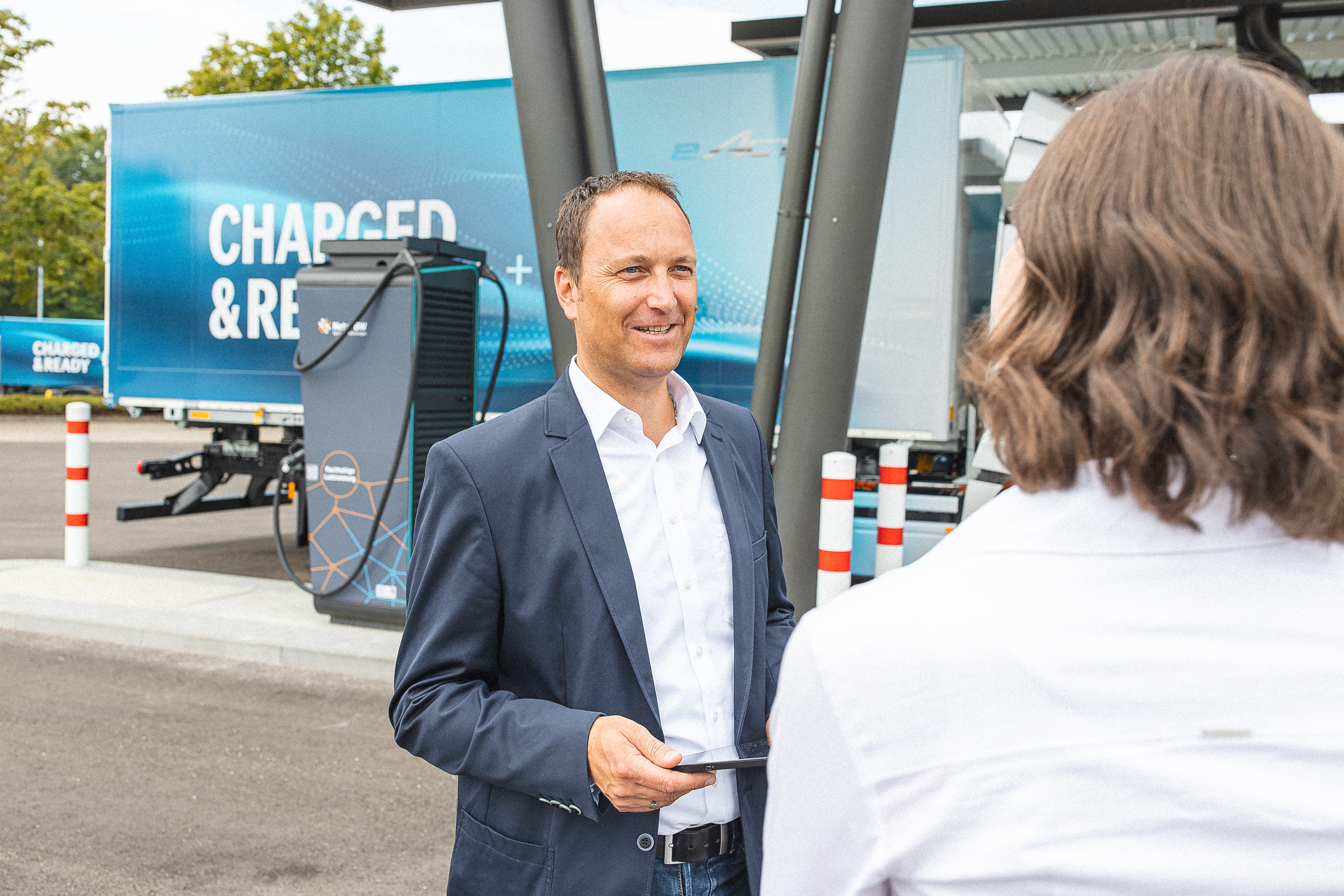 Lächelnder Mann im Anzug vor Ladesäule für E-LKW.