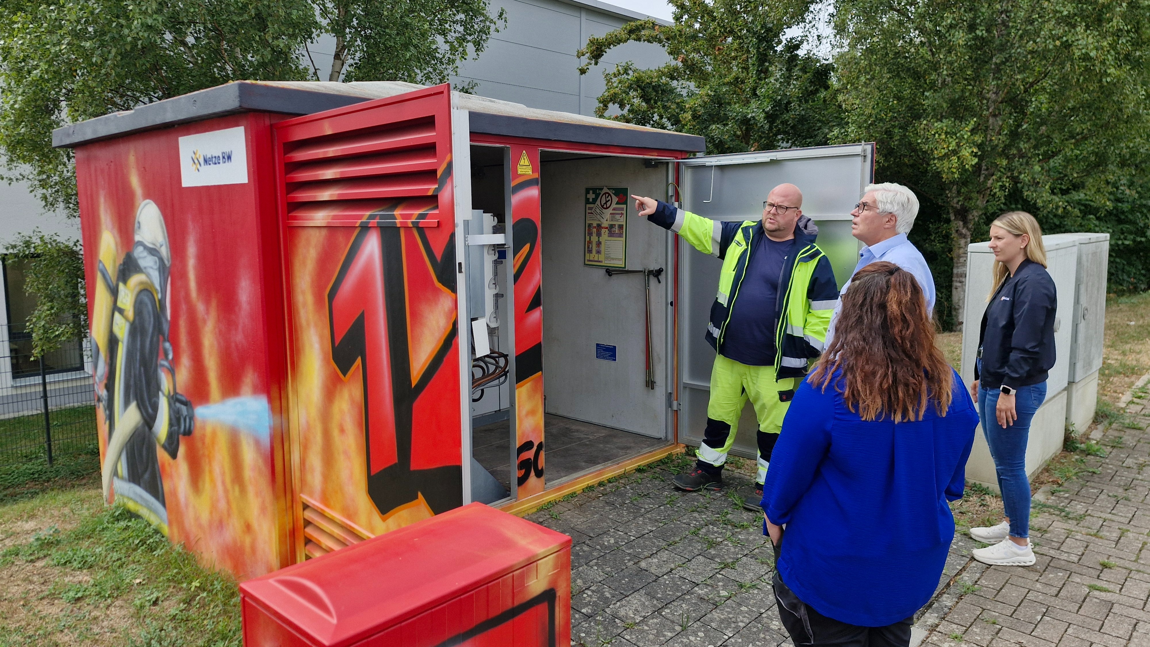 Vor geöffneten Türen der neu gestalteten Trafostation: Dennis Baumann (Teamleiter Netzbetrieb der Netzregion Kraichgau) zeigt 3 Personen die komplexe Technik im Innenraum der Trafostation