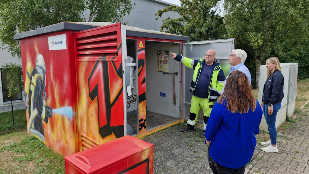 Vor geöffneten Türen der neu gestalteten Trafostation: Dennis Baumann (Teamleiter Netzbetrieb der Netzregion Kraichgau) zeigt 3 Personen die komplexe Technik im Innenraum der Trafostation
