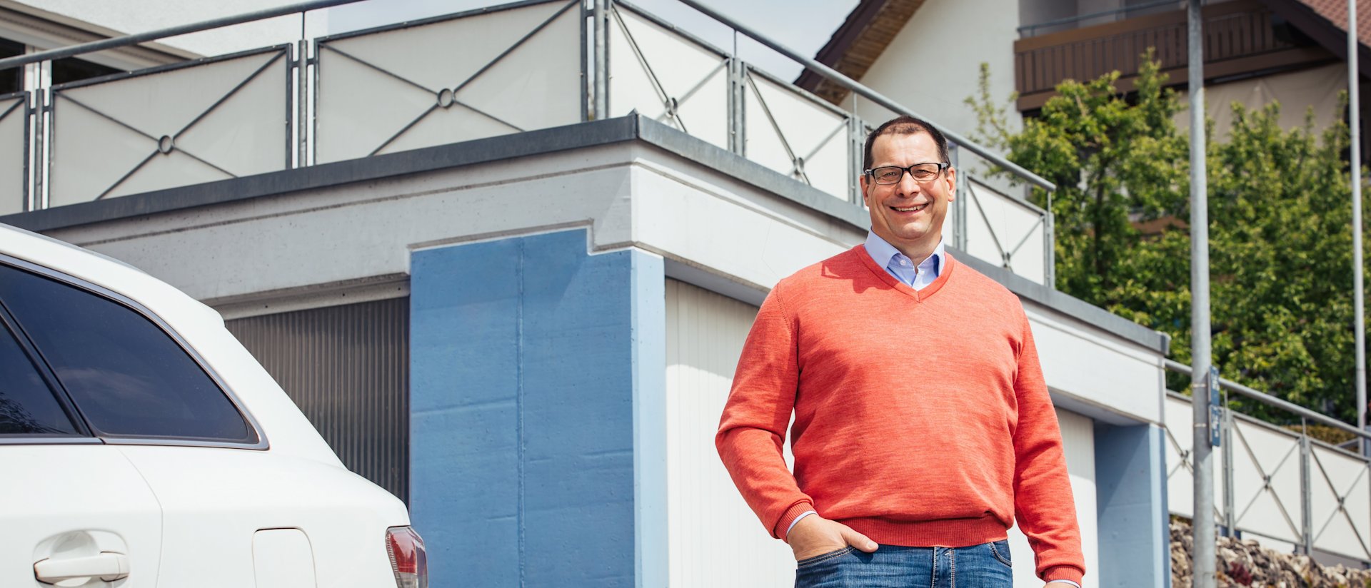 Mann in orangem Pullover steht vor Haus mit Garage.