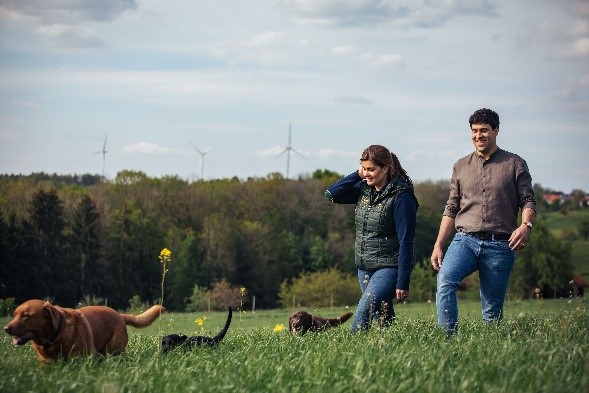 Mann und Frau mit Hunden auf Wiese, Windräder im Hintergrund.