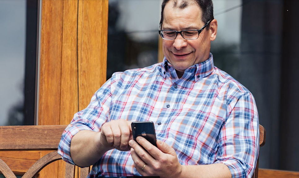 Lächelnder Mann mit Brille benutzt Smartphone vorm Haus sitzend.